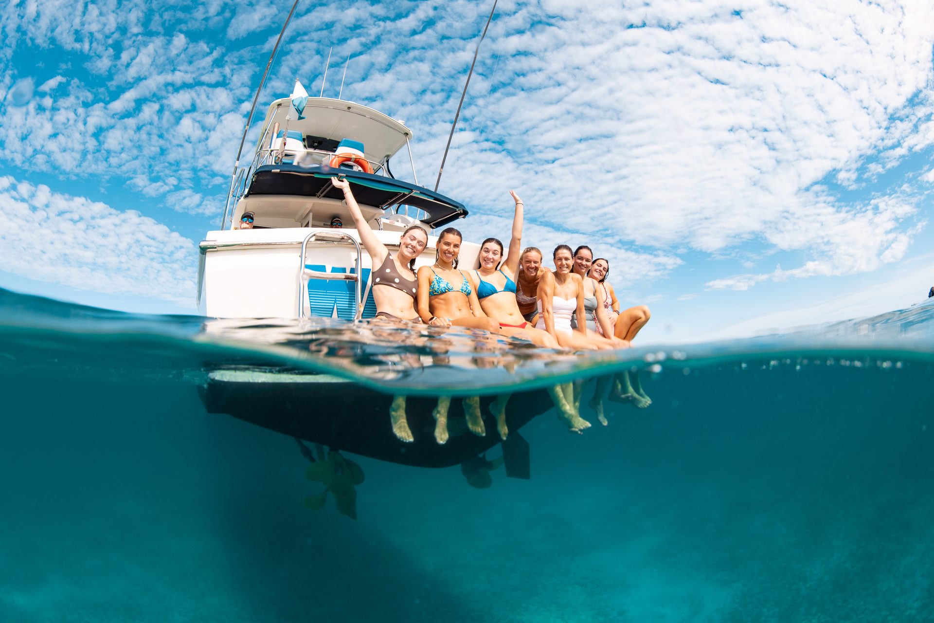 There are seven women on the back deck of Wave Rider, Live Ningaloo's boat. They are all smiling, and two people have their hands in the air in celebration. The image has blue sky with white fluffy clouds and the water of the Ningaloo reef below. You can see their feet too.